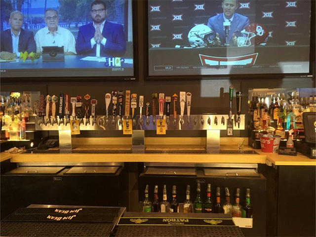 Bar counter with various beer taps and liquor bottles below two mounted TV screens showing sports programs.