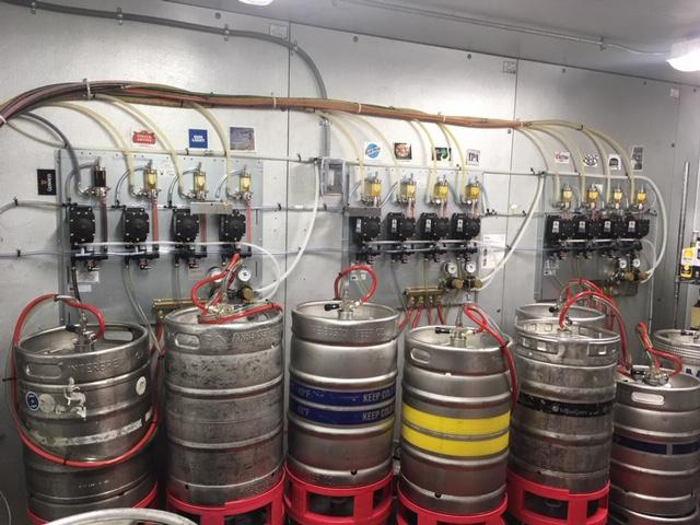 A row of beer kegs connected to a complex system of tubes and valves in a brewery or bar storage room.