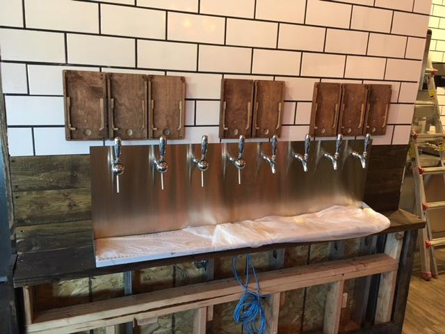 A wall with six metal beverage taps below wooden plaques, set against white tile and wooden panels.