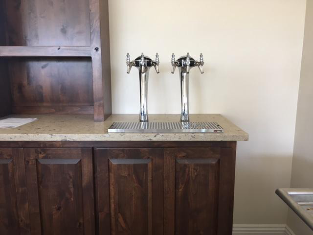 Two stainless steel beer taps on a countertop with wooden cabinetry below, set against a beige wall.