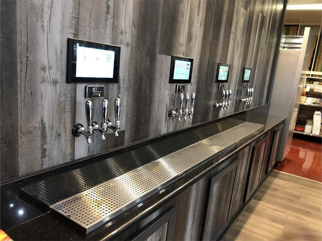 A row of self-service beer taps with digital screens on a wooden wall surface.
