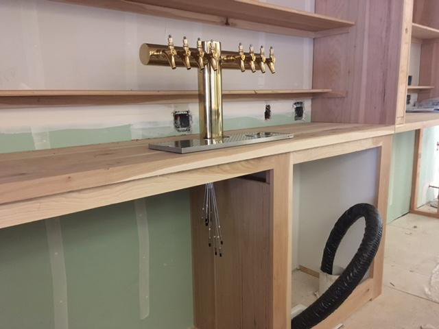 Unfinished wooden bar counter with a gold beer tap featuring eight spouts, surrounded by shelves and unpainted walls.