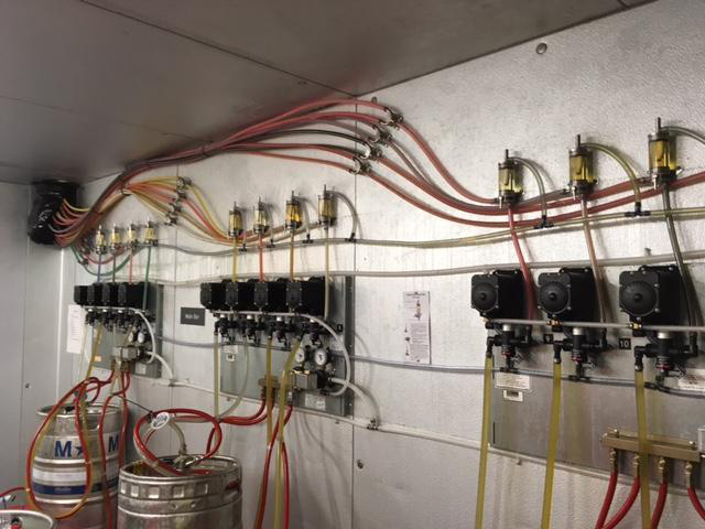 Beer dispensing system with red and yellow tubes connected to a row of regulators, attached to a wall. A metal keg is visible below.