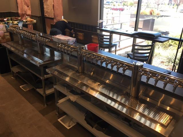 Inside a bar, two long rows of beer taps on metal counters are set up near windows with stools. A person is working in the background.