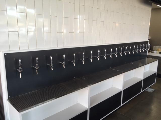 A long row of beverage taps is mounted on a dark panel against a wall with white tiles. Below the taps, there are empty shelves and a grated counter surface.